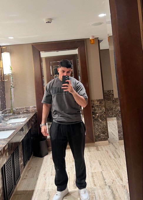 A mirror selfie of a young man wearing a gray 'Notre Dame' sweatshirt, black sweatpants, and white sneakers in what appears to be an upscale bathroom or hotel lobby.