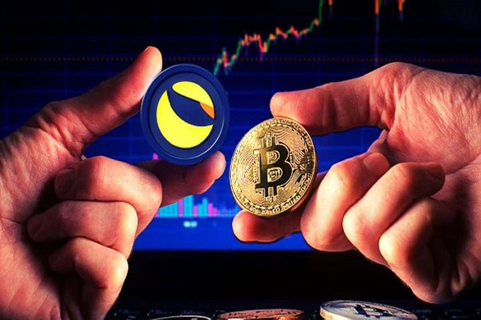 Two hands hold up crypto coins—one a blue Terra/Luna (LUNC) token and the other a physical Bitcoin—against a backdrop of candlestick price charts, symbolizing a diamond-hands stance of holding $LUNC through market volatility.