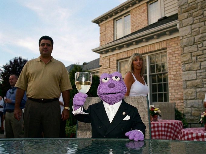 The image features a purple puppet sitting at an outdoor table, holding a wine glass. The puppet appears to be dressed in a suit and tie, giving it a sophisticated appearance. In the background, there are people present who seem to be enjoying their time together. A man is seen wearing a yellow shirt while another person is wearing a blue shirt. There's also a woman standing nearby with her back turned towards the puppet. The scene takes place in front of a house and appears to be set up for an outdoor gathering or event, possibly a party or social function.