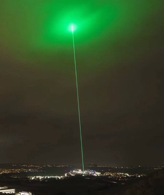 A bright green laser beam shoots vertically into the night sky above a city lit with scattered lights.