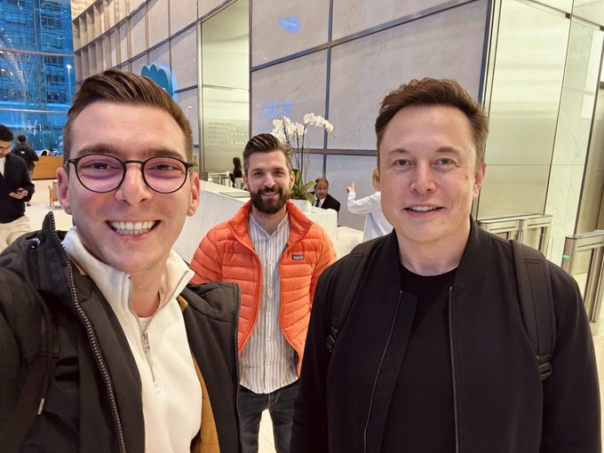 Photo of three men, including one resembling Elon Musk, taking a selfie in what appears to be a lobby or event venue.