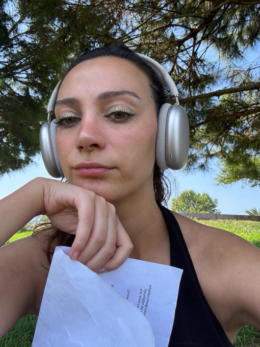 A woman wearing silver over-ear headphones sits outdoors under pine trees, resting her chin on her hand while holding a piece of paper with handwritten text that includes the words 'RIGHT', 'need any', and 'professional'.
