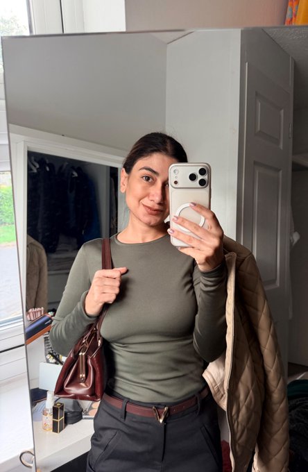 A mirror selfie of a woman holding a smartphone, wearing an olive green long-sleeved fitted shirt, gray trousers, and a brown belt with a gold buckle, while carrying a brown leather handbag and a beige quilted coat.