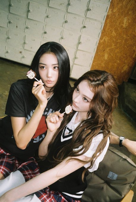Two young women with long dark hair pose together indoors, each holding a pink lollipop near their face, with a wall of small drawers behind them.