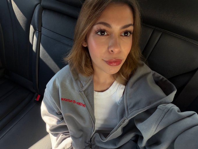 A woman with shoulder-length brown hair taking a selfie in a car, wearing a gray zip-up hoodie over a white top.