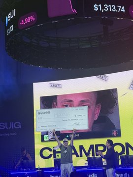 Two winners celebrate on stage with a giant $25,000 check at the first Sui trading competition, with the event screen displaying "@Tradermayne" alongside a 4.99% gain and a $1,313.74 price figure for the SUISUI token. The celebratory moment captures the champion's victory amid the competition's branding visible in the background.