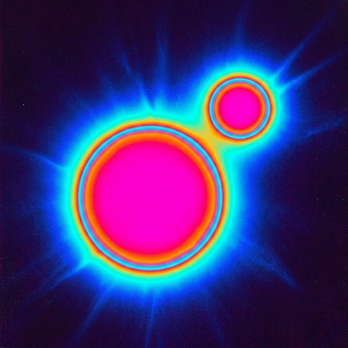 An abstract image showing two glowing pink spheres with concentric colorful rings of blue, yellow, and orange, surrounded by a radiant blue aura against a dark background.