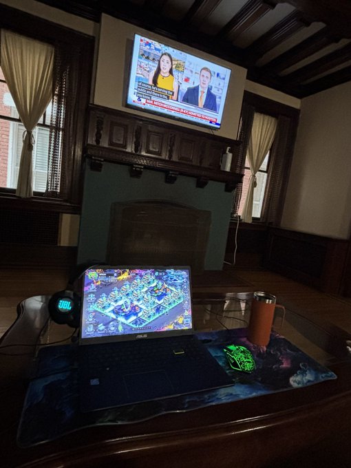 The image shows a cozy living room setting featuring a wooden fireplace, a television mounted above a fireplace, and a laptop placed on top of a coffee table or desk. A mouse can be seen next to the laptop, indicating that someone might have been working or gaming at this location. There is also an orange cup sitting on the table near the laptop. The room appears to be well-lit with natural light coming in from two windows and artificial lighting provided by the television.