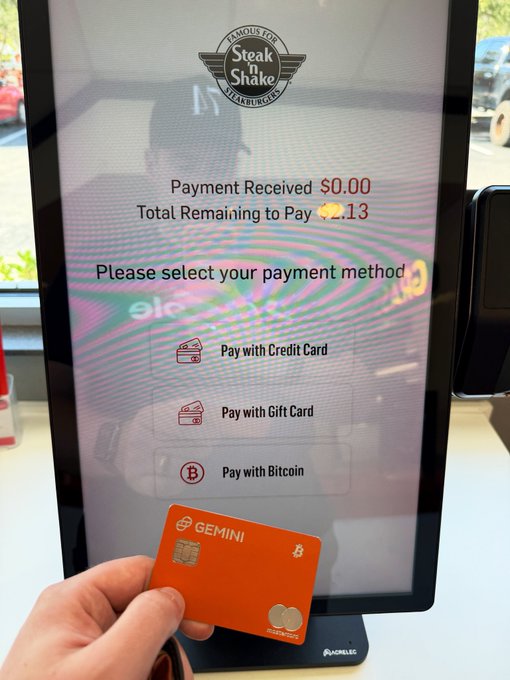 A Steak 'n Shake kiosk payment screen showing 'Payment Received $0.00' and 'Total Remaining to Pay $?.13', with a hand holding an orange Gemini Bitcoin gift card in the foreground.