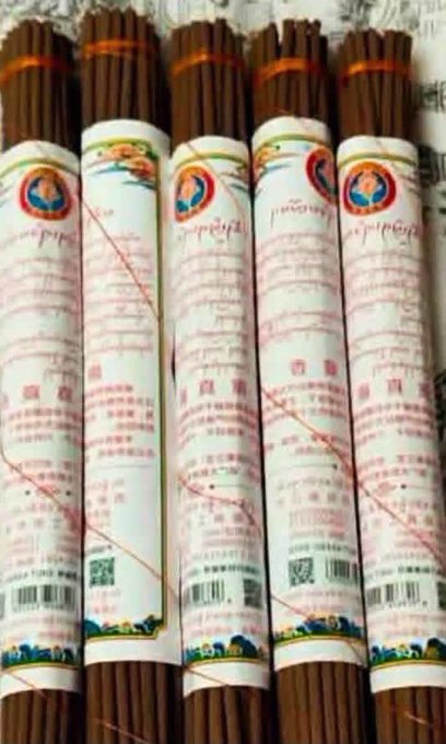Several bundles of incense sticks with labels featuring Tibetan or Chinese characters, barcodes, and colorful circular logos are lined up closely together.