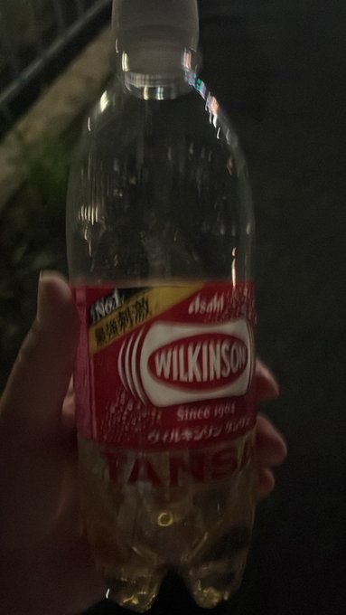 A hand holding a plastic bottle of Wilkinson Tansan sparkling water at night, with a red label reading 'WILKINSON' and 'Since 1904'.