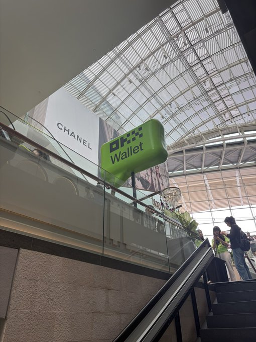 A shopping mall interior features a green OKX Wallet inflatable display and a Chanel storefront banner, with shoppers riding an escalator beneath a large skylight.