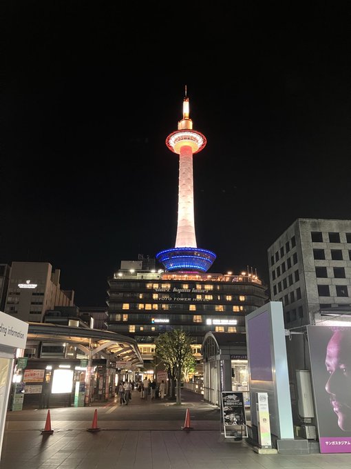 A nighttime view of the illuminated Kyoto Tower, glowing red, white, and blue, towering over surrounding buildings in an urban plaza.