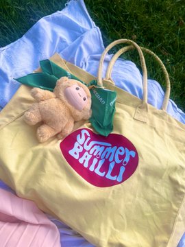 A 'Summer Brilli' tote bag in pale yellow with a pink and blue logo lies on a blue cloth on grass, accompanied by a small stuffed monkey toy with a green bow.