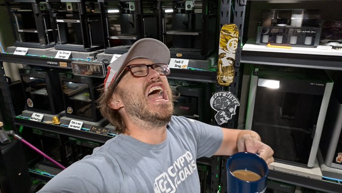 A man wearing a 'CRYPTO JEANS' t-shirt and a cap, laughing enthusiastically in a workshop or storage room filled with shelves of Bitcoin mining hardware, holding a blue mug.