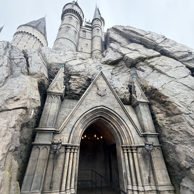 A gothic-style stone castle entrance features a pointed archway doorway built into a rocky cliff face, with tall spired towers visible at the top and lanterns mounted beside the door.