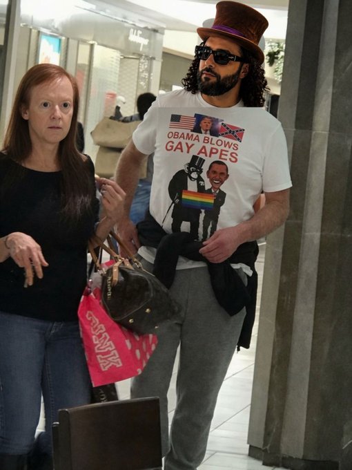 A man wearing a top hat, sunglasses, and a t-shirt reading 'OBAMA BLOWS GAY APES' stands in a building lobby next to a woman with a handbag.
