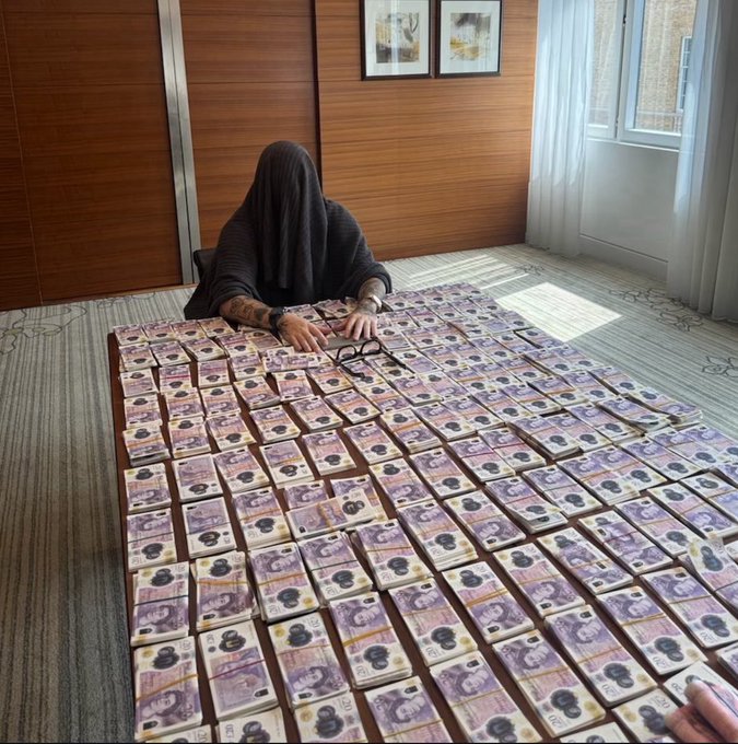 A person whose face is covered by dark fabric sits at a table covered with stacks of currency notes in various denominations.