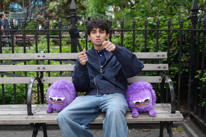 A young man in a navy bomber jacket and jeans sits on a wooden park bench between two purple furry plush toys, holding a microphone and gesturing while speaking.