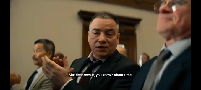 A still from a film or TV show showing a man in a dark suit clapping, with the subtitle reading 'She deserves it, you know? About time.'