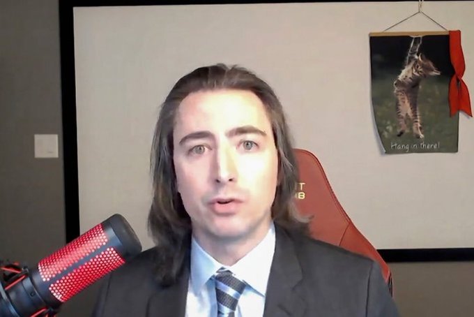 A man in a suit and tie speaks into a red microphone in what appears to be a recording or streaming setup, with a "Hang in there!" kitten poster visible behind him on the wall.