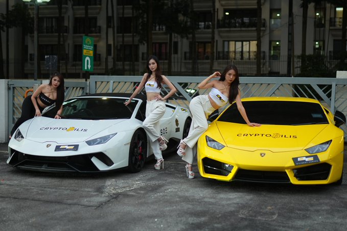 Photo of three women posing with two Lamborghini Huracán cars branded with 'CryptoBilis' logos — one white car with number 606 and one yellow car — parked at night outside a building.