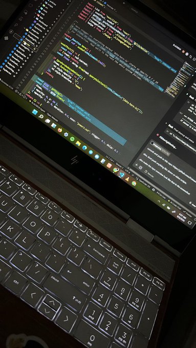 A laptop displaying a code editor with colorful syntax highlighting on the screen, alongside a Discord or messaging application in the background, with a black keyboard visible in the foreground.