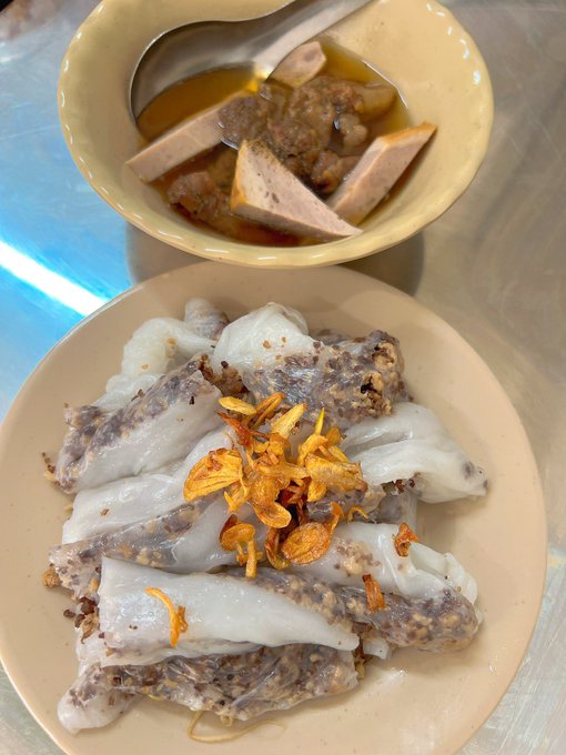 Two dishes of food on a metal table - one plate of rolled rice noodle sheets topped with fried shallots and a bowl of broth with sliced meat.