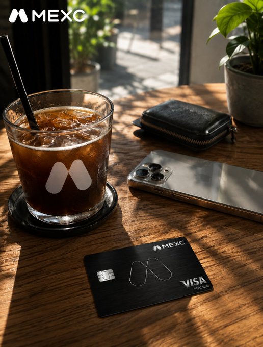 A product photograph showing a MEXC-branded iced coffee, a smartphone, a black leather wallet, and a MEXC Visa Platinum card arranged on a wooden café table with plants in the background.