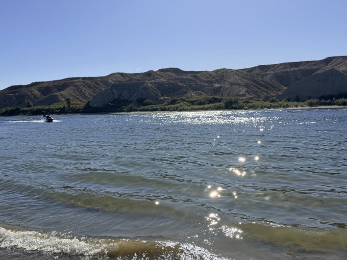 A scenic view of a wide blue river with sunlight reflecting off the surface, a person jet skiing, and arid mountains in the background under a clear sky.