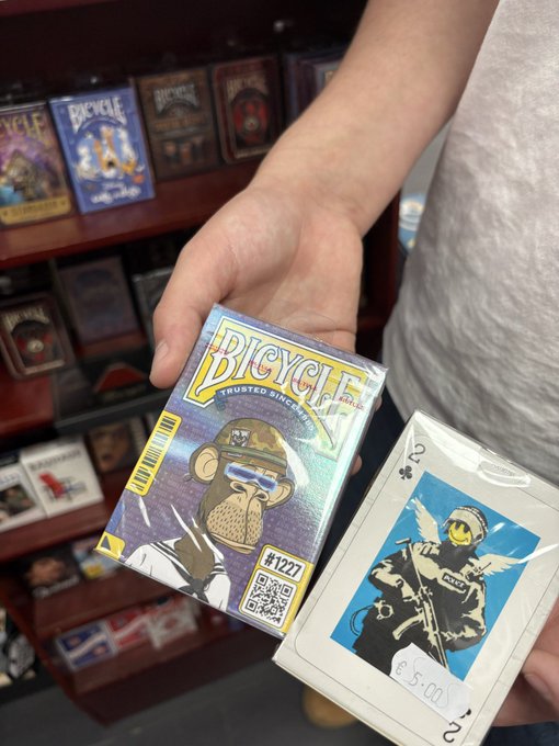 A hand holds up two sealed 'Bicycle' brand playing card decks (No. 1227) featuring artwork of a monkey in sunglasses, priced at €5.00, in what appears to be a magic shop.