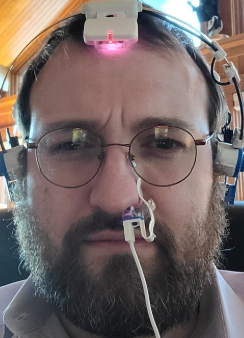 A bearded man wearing round glasses and a head-mounted device with a red glowing light looks directly at the camera.