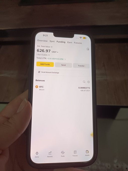 A hand holding a smartphone displaying a cryptocurrency trading app showing an estimated total value of 626.97 USDT with 0.00993773 BTC in balances.
