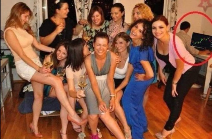 A humorous photo of a group of women posing together at a party, with a man circled in red in the background sitting at a desk looking at a trading chart on his monitor.