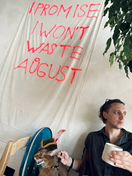 A person sitting next to a taxidermy cat wearing a hat, beneath a cloth banner with handwritten red text reading 'I PROMISE I WON'T WASTE AUGUST'.