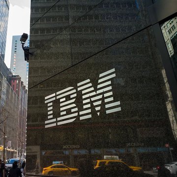 The image features a tall building with the IBM logo prominently displayed, along with the words "IBM Steller" written below it. Below the building, there are two yellow taxis driving down the street, one of which has its headlights on. A few people can be seen walking around in the area as well.