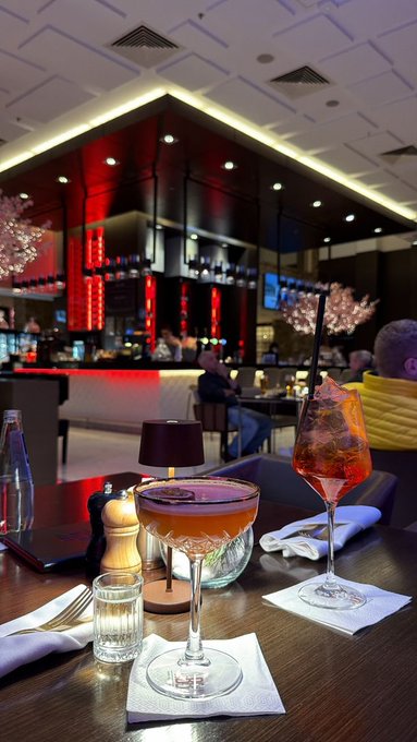 Two cocktails—one an orange aperitif-style drink and the other a tall glass with ice and a black straw—sit on a wooden table in an upscale bar or restaurant with red ambient lighting.