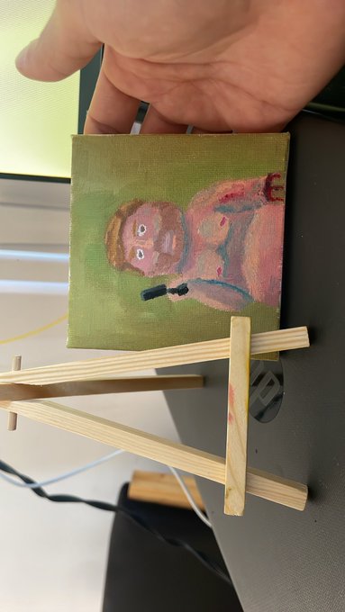 A hand holding a small canvas painting of a figure in a pink/red hoodie on a green background, resting on a wooden display easel.