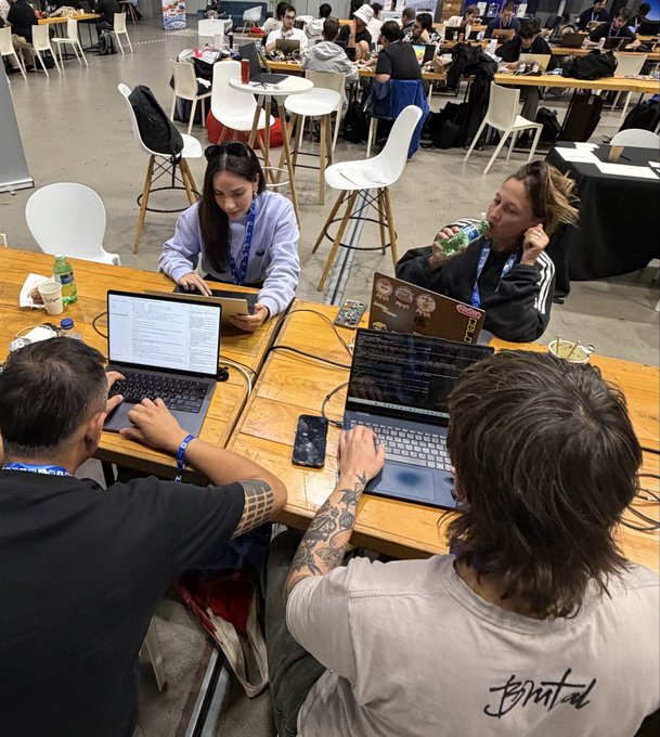 A group of four people working on laptops at a wooden table in what appears to be a hackathon or co-working space, with other participants visible in the background.
