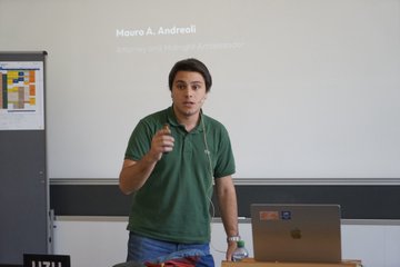 A man in a green polo shirt stands in front of a projector screen displaying "Mauro A. Andreoli, Behörden und Hörigkeit Appreciator" while holding a microphone during what appears to be a lecture. The image accompanies a post expressing gratitude for returning to teach at the University of Zurich, reflecting his passion for mentoring and sharing knowledge in an academic setting.