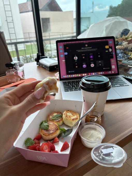 A person holds a piece of pancake above a pink box containing small pancakes, strawberries, and mint, alongside a coffee cup and cream container, with a laptop open on the table behind.