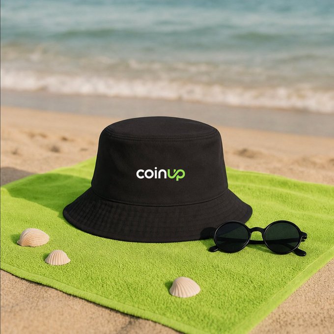 The image features a black bucket hat laying on a green towel at the beach. There are also sunglasses placed next to the hat, suggesting that someone might be spending time outdoors enjoying the sun and sand. The photo has been captioned with "GM CoinUp fam A new week, just in time for Children's Day May everyone bring a bit of childlike wonder on-chain today, with less anxiety and more curiosity #CoinUp".