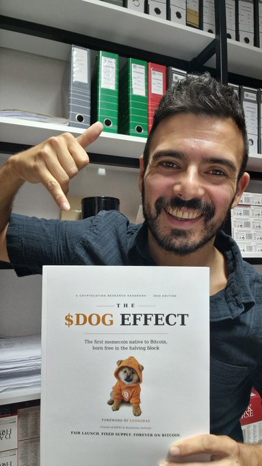 A smiling man with a beard and finger guns holds up the book 'The $DOG EFFECT: The first memecoin native to Bitcoin, born free in the halving block,' with a Dogecoin dog mascot on the cover.