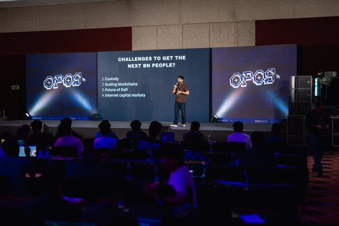 A speaker presents at the Solana Summit India in Mumbai, addressing an audience of 700+ attendees about "Challenges to Get the Next Bn People?" — listing Custody, Scaling blockchains, Future of DeFi, and Internet capital markets — while flanked by QPoS-branded screens in a large conference hall.