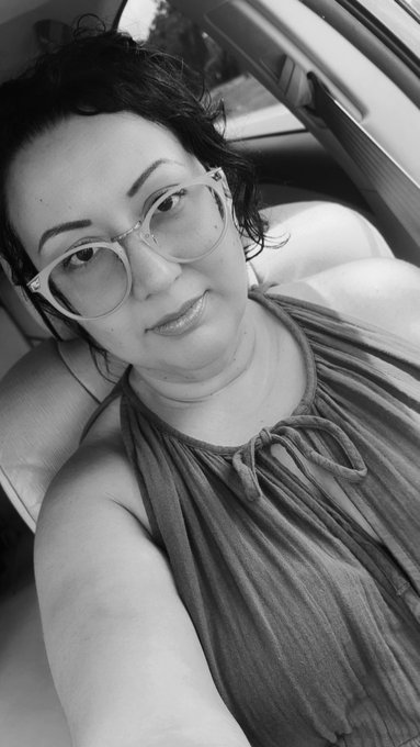 A black-and-white selfie of a woman wearing clear-framed glasses and a halter top, sitting inside a car.