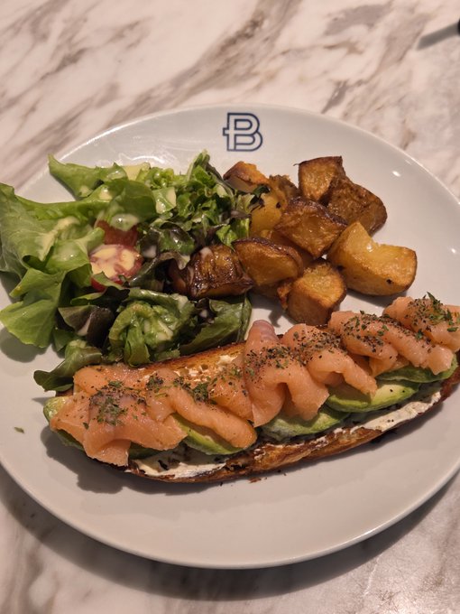 A plate branded with a blue "B" features smoked salmon draped over avocado toast, accompanied by golden roasted potatoes and a fresh green salad. The "Minted and gone in minutes" caption playfully suggests the delicious meal was devoured almost instantly, befitting the "B" branding likely of a café or restaurant.