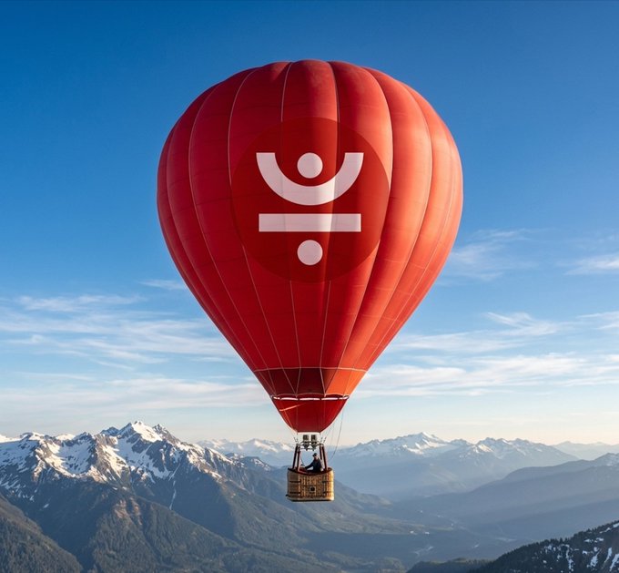 A red hot air balloon with a white logo floats above snow-capped mountains against a blue sky.