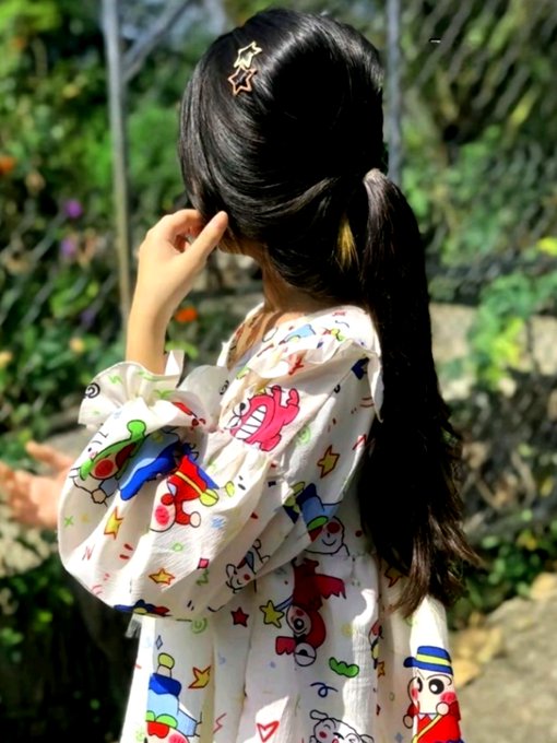 A woman with long dark hair tied in a ponytail with star-shaped clips poses outdoors in a garden, wearing a white sheer dress with colorful cartoon character prints.