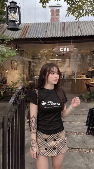 A young woman with long dark hair stands outside a café wearing a black t-shirt printed with "AXIS ROBOTICS THE FUTURE IS ROBOTICS" and a plaid skirt.
