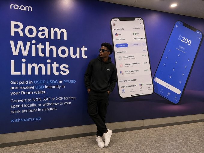 A man posing in front of a Roam 'Roam Without Limits' advertisement billboard promoting a crypto wallet app with $55,000.00 and $10,000,000.00 USD balances, and a $200 payment screen.